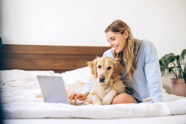 Evden uzakta çalışan mutlu serbest çalışan kadın, yatağında dizüstü bilgisayar kullanarak dost canlısı Golden Retriever köpeğiyle eğleniyor, rahat ve rahat bir atmosfer yaratıyor.