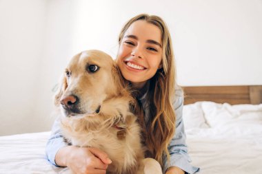 Gülümseyen genç bir kadın, bir yatakta uzanırken Golden Retriever köpeğini kucaklıyor, sıcak bir ev ortamında şefkat ve arkadaşlığın tadını çıkarıyor.