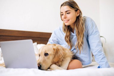 Genç bir kadın evde çalışıyor ya da ders çalışıyor. Yanında Golden Retriever köpeği ile yatakta rahat bir şekilde oturuyor. Dizüstü bilgisayar kullanıyor.