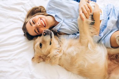 Mutlu genç bir kadın, açık mavi pijama giymiş, neşeli bir şekilde köpeğine sarılıyor, kahkaha ve şefkatle dolu neşeli bir sabahın tadını çıkarıyor.