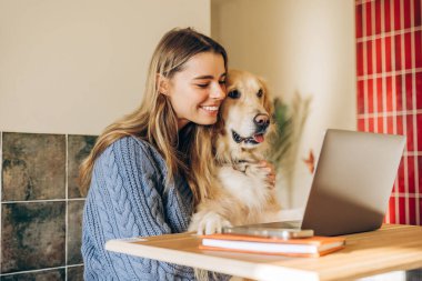 Gülümseyen, serbest çalışan genç bir kadın evinde dizüstü bilgisayar başında çalışırken Golden Retriever köpeğine sarılıyor, uzaktan çalışmanın ve evcil hayvan arkadaşlığının keyfini çıkarıyor.