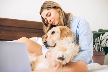 Genç kadın serbest çalışan, Golden Retriever köpeğini kucaklarken uzaktan çalışıyor. Ev ortamında rahat ve üretken bir iş ortamı yaratıyor.