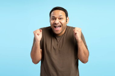 Hevesli adam sevinç ve zaferini yumruklarını kaldırarak ve ağzı açık bir şekilde canlı bir mavi zemine karşı zafer ya da başarıyı kutlayarak ifade ediyor.