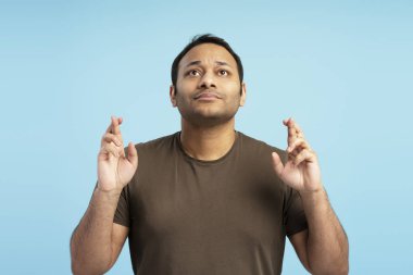 Umut dolu yetişkin bir adam parmaklarını çapraz yapıp yukarı bakıyor, sakin mavi bir arka plana karşı olumlu bir şey olmasını diliyor, beklenti ve arzuyu somutlaştırıyor.