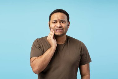 Güçlü diş ağrısı olan orta yaşlı bir adamın stüdyo portresi acı dolu bir ifadeyle yanağına dokunuyor, mavi arka planda izole edilmiş, sağlık ve diş sağlığı için mükemmel. Diş sağlığı konsepti