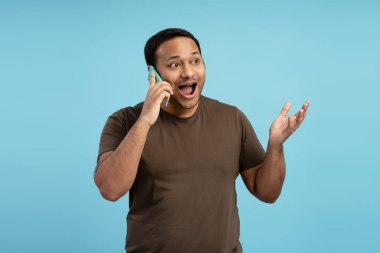 Heyecanlı bir adam cep telefonuyla konuşurken, canlı mavi bir arkaplana karşı şaşkınlık ve coşkuyu ifade ederken eliyle işaret ediyor. Çevrimiçi iletişim kavramı