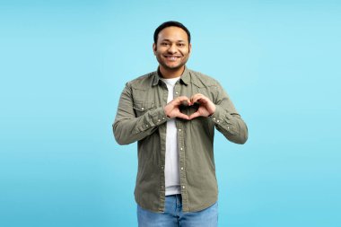 Dost canlısı, orta yaşlı, kalp şeklini elleriyle gösteren, sevgisini ve özenini ifade eden, kameraya gülümseyen, günlük kıyafetler giyen, mavi stüdyo arka planında soyutlanmış yetişkin bir adam. Aşk konsepti