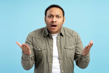 Şok olmuş sıradan bir adamın, kafası karışmış, ağzı açık, avuçları açık, mavi arka planda şaşkınlık ve şaşkınlık ifadesi gösteren bir portresi.