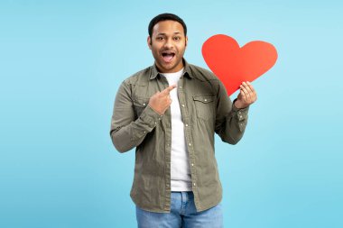Şaşkın, koyu renk saçlı, sıradan elbiseli bir adam elinde tuttuğu büyük kırmızı bir kalbe parmağıyla işaret ediyor, açık mavi bir arkaplana karşı duruyor. Reklam kavramı