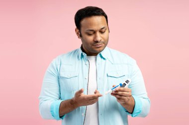 Konsantre Hintli adam şeker hastalığı testi için glukometre ve tükenmez kalem kullanıyor. Evde glikoz seviyesini kontrol ediyor. Pembe arka planda duruyor. Sağlık hizmeti kavramı