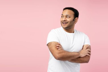 Kollarını çapraz bağlamış, beyaz tişört giymiş, pembe bir arka planda gülümseyen, mutluluk ve kendine güveni ifade eden neşeli orta yaşlı Hintli bir adamın stüdyo portresi. Alışveriş konsepti