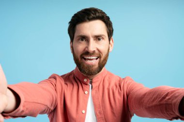Stüdyoda mavi arka planda selfie çeken mutlu genç sakallı adamın portresi. Alışveriş, reklam konsepti.