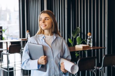 Genç bayan mimar elinde dizüstü bilgisayar ve planlar ile modern mobilya ve parlak doğal ışıkla dolu bir ofise bakarken gülümsüyor.