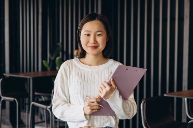 Kendine güvenen Asyalı genç iş kadını elinde bir pano ve kalem tutarken gülümsüyor, geçmişi bulanık, profesyonellik fışkıran modern bir ofis alanında duruyor. İş konsepti