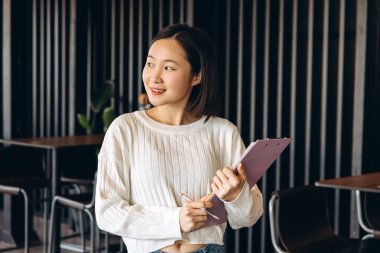 Genç Asyalı kadın, öğrenci ya da girişimci, modern uzayda pano ve kalemle duruyor, düşünceli bir şekilde yan tarafa bakıyor, muhtemelen bir sonraki görevini ya da fikrini düşünüyor. Başarılı iş konsepti