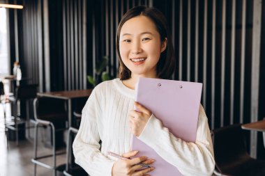 Asyalı genç bir iş kadınının portresi elinde bir dosya ve kalem, modern bir ofis alanında duruyor, kurumsal bir ortamda güven ve profesyonelliği öneriyor. İş konsepti