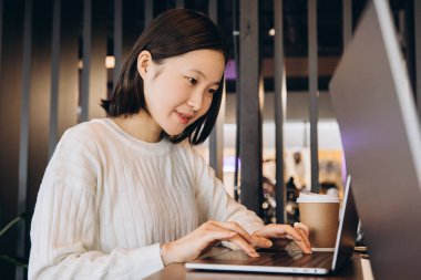 Laptopuna odaklanan Asyalı genç bir kadın, modern bir çalışma alanındaki bir masada özenle çalışarak rahat ve rahat bir ortamda kahvesini yudumluyor. Çevrimiçi teknoloji kavramı