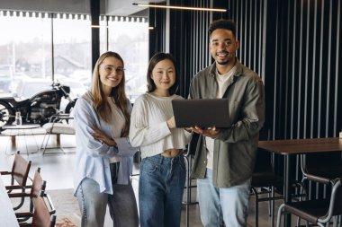 Çeşitliliği temsil eden üç genç profesyonel, çağdaş bir ofis alanında dizüstü bilgisayar kullanarak takım çalışması ve yenilik sergileyerek birlikte çalışıyorlar.