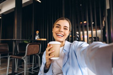 Neşeli iş kadını, modern kafe molasında selfie çekerken ve kahve fincanını tutarken geniş bir gülümseme sergiliyor, rahatlama ve bağlanma anının tadını çıkarıyor. Çevrimiçi teknoloji kavramı