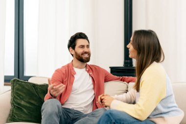 Genç adam ve kadın rahat bir kanepede oturuyor, keyifli bir sohbete dalıyorlar, modern oturma odalarında sıcak ve samimi bir ortam yaratıyorlar.