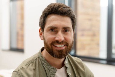 Gülümseyen, sakallı bir iş adamının portresi gündelik bir gömlek giyiyor, modern ofis ortamında kendine güven ve profesyonellik yayıyor, başarıyı ve yaklaşımı somutlaştırıyor.