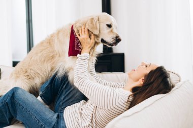 Genç bir kadın rahat beyaz bir kanepede uzanmış, Golden Retriever köpeğiyle şakalaşıyor, kırmızı bir bandana takıyor, evinde rahatlatıcı bir anın tadını çıkarıyor.