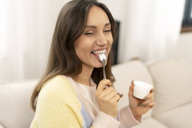 Gülümseyen kadın, oturma odasındaki rahat bir kanepede dinlenirken bir kaşık yoğurdun tadını çıkarıyor.