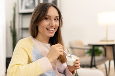 Gülümseyen kadın rahat bir oturma odasında koltukta dinlenirken yoğurdun tadını çıkarıyor, sağlıklı beslenme alışkanlıklarını ve refah ve keyif dolu rahat bir yaşam tarzını somutlaştırıyor.