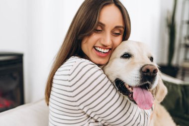 Gülümseyen genç kadın, evdeki oturma odasındaki kanepede otururken sevimli Golden Retriever köpeğini kucaklıyor. Samimi bir ev ortamında sevgi, ilgi ve mutluluk gösteriyor.