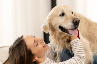 Genç bir kadın kanepede uzanıyor, sevgili golden retriever 'ına sarılıyor, evdeki eğlenceli etkileşimlerden zevk alırken neşe ve şefkat anlarını paylaşıyor.