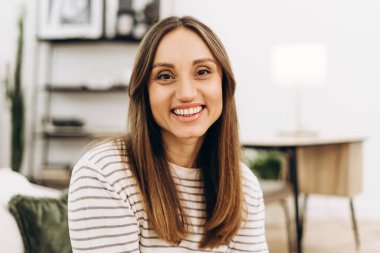 Uzun kahverengi saçlı, çizgili gömlek giyen, rahat oturma odasında dinlenirken geniş bir gülümsemeyle gülümseyen, evinde huzur ve mutluluğun tadını çıkaran neşeli bir genç kadın portresi.