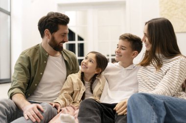 Dört kişilik aile evde birlikte vakit geçiriyorlar, rahat bir şekilde oturup hoş bir sohbete dalıyorlar, sıcak ve sevgi dolu bir ortam yaratıyorlar.