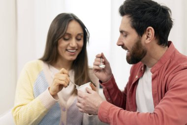 Gülümseyen çift evde yoğurtlarını paylaşıyor, birlikte sağlıklı bir atıştırmalık yiyor ve rahat yaşam alanlarında bağlantının ve mutluluğun tadını çıkarıyorlar.