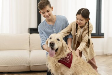 Gülümseyen erkek ve kız kardeşler, parlak kırmızı bir bandana takmış sevimli Golden Retriever köpeklerini okşayarak, sıcak oturma odasında birlikte otururken, neşe ve şefkat dolu bir anın tadını çıkarıyorlar.