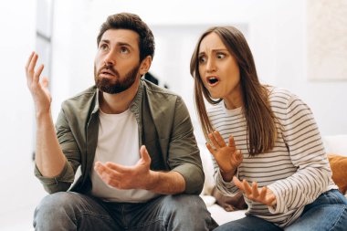 Evde tartışan genç bir çift. Elleriyle hareket ediyorlar. Sinirli ve sinirli görünüyorlar.