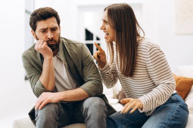 Genç bir kadın evdeki kanepede oturan üzgün kocasına bağırıyor ve tartışıyor. İstifa etmiş görünüyor ve sakalına eliyle dokunuyor.