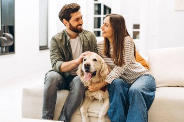 Genç çift evlerindeki kanepede birlikte vakit geçiriyorlar. Sevimli Golden Retriever köpeklerini okşayıp birbirlerine gülümsüyorlar. Barışçıl bir bağ ve şefkat anının tadını çıkarıyorlar.