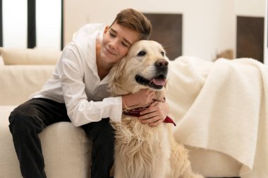 Oturma odasında rahat bir kanepede otururken Golden Retriever köpeğini kucaklayan mutlu genç, evde bir anlık şefkat ve arkadaşlığın tadını çıkarıyor.