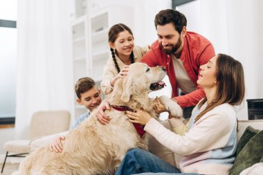 Gülümseyen aile üyeleri, oturma odasında rahat bir kanepede oturup sevgi ve bağlılık dolu bir anın tadını çıkarırken, sevimli Golden Retriever köpeklerini seviyorlar.
