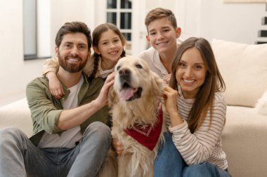 Gülümseyen ebeveynler ve çocuklar, oturma odasında oturmuş sıcak bir aile anının tadını çıkarırken, dost canlısı Golden Retriever köpeklerine sarılıyorlar.