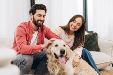 Gülümseyen bir çift, modern bir oturma odasında rahat bir kanepede rahat rahat otururken dost canlısı golden retriever 'larını okşayarak sıcak bir ev ortamında kaliteli zaman geçiriyorlar.