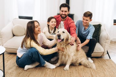 Neşeli bir aile, ebeveynler ve çocuklar, oturma odasında yerde ve kanepede oturuyor, dost canlısı Golden Retriever köpeklerini okşuyor, birlikte kaliteli zaman geçiriyorlar.