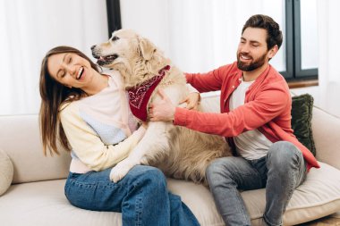 Genç ve gülümseyen bir çift, evdeki oturma odasında rahat bir kanepede otururken bandana takan sevimli Golden Retriever köpeğiyle oynuyorlar.
