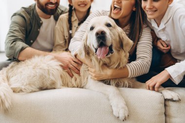 Gülümseyen aile sevgili Golden Retriever köpeğini kucaklarken rahat bir kanepede, samimi evlerinde sevgi ve bağlılık dolu bir anın tadını çıkarıyorlar.