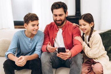 Mutlu bir aile evde eğleniyor, baba gülümseyen çocuklarla komik bir video paylaşıyor, onu kucaklıyor, neşeli anları ve güçlü aile bağlarını yakalıyor.