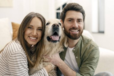 Genç, gülümseyen bir çift oturma odasında yerde oturmuş, sevimli Golden Retriever köpeklerine sarılıyor ve sıcak bir anın tadını çıkarıyorlar.