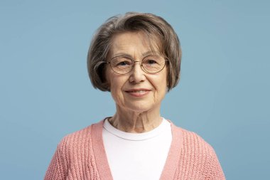 Gözlüklü, kendine güvenen ve mavi arka planda poz veren mutlu yaşlı kadının stüdyo portresi.