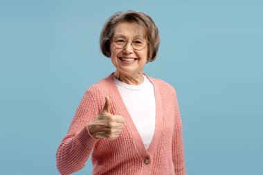 Neşeli, yaşlı bir kadın, mavi arka planda olumlu ve onaylı olduğunu belirterek baş parmak işareti yapıyor ve gülümsüyor.