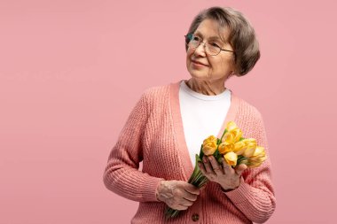 Gözlüklü, pembe hırkalı, elinde pembe bir demet sarı laleyle pembe arka planda kameradan başka tarafa bakan mutlu bir büyükanne. Kutlama konsepti
