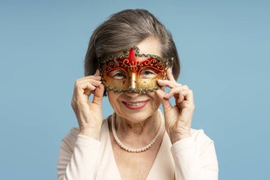 Venedik karnaval maskesi takan, gülümseyen ve Mardi Gras partisine hazırlanan zarif yaşlı kadının stüdyo portresi.